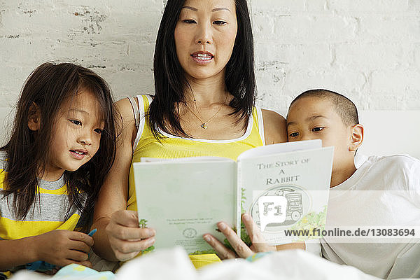 Mother reading book to children while sitting on bed at home