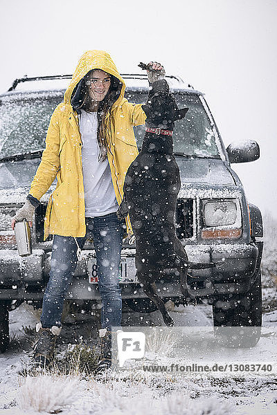 Woman playing with dog while standing off-road vehicle on field during winter