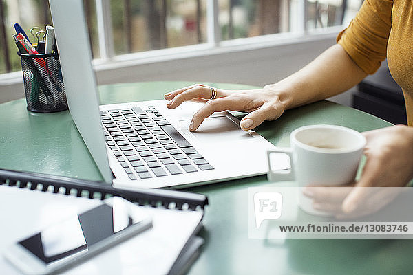 Cropped image of businesswoman holding coffee cup while using laptop in office