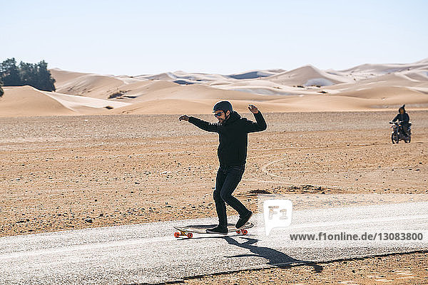 Skateboard fahrender Mann in voller Länge auf der Straße inmitten des Feldes gegen den Himmel bei Sonnenschein