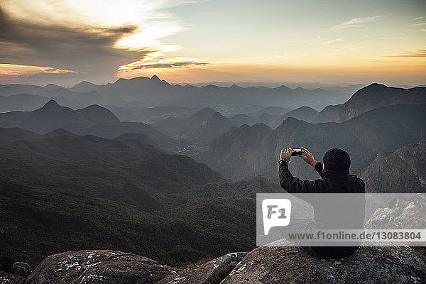 Rear view of hiker photographing through smart phone while sitting mountain during sunset