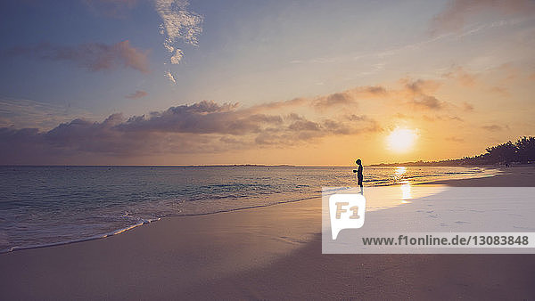 Side view of silhouette boy standing on shore at beach against sky during sunset