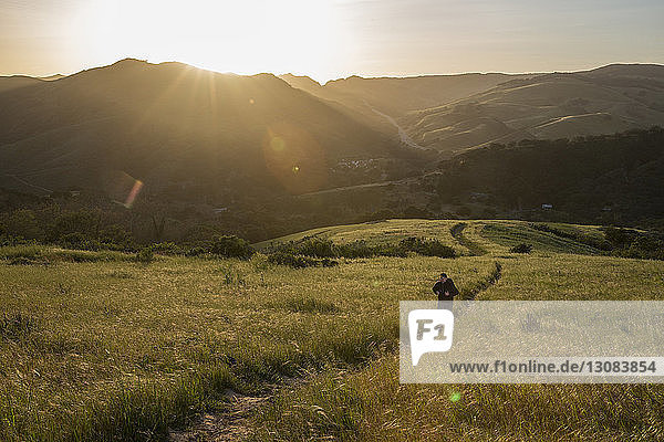 Hochwinkelaufnahme eines Mannes auf einem Wanderweg inmitten eines Grasfeldes gegen Berge bei Sonnenuntergang