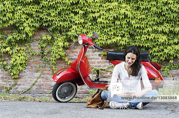 Woman using mobile phone while sitting against motor scooter and ivy wall