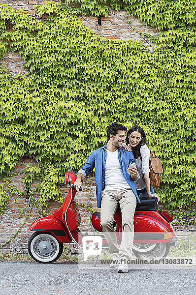 Couple using smart phone while standing with motor scooter against ivy wall