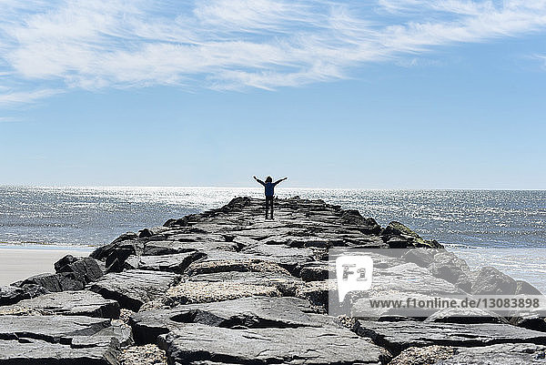 Rear view of girl with arms outstretched standing at rocky beach against sky during sunny day