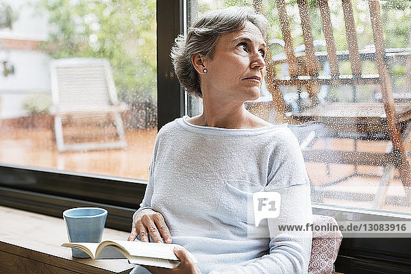 Nachdenkliche reife Frau  die durch ein Fenster schaut  während sie zu Hause ein Buch in der Hand hält