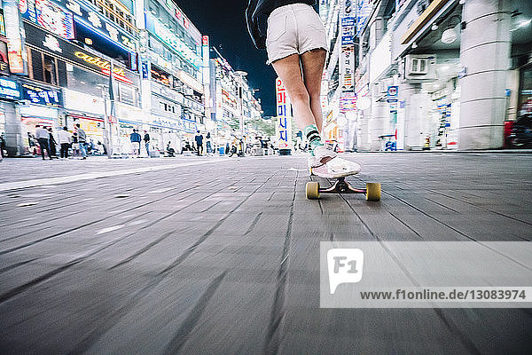 Low section of woman skateboarding on footpath in illuminated city during night