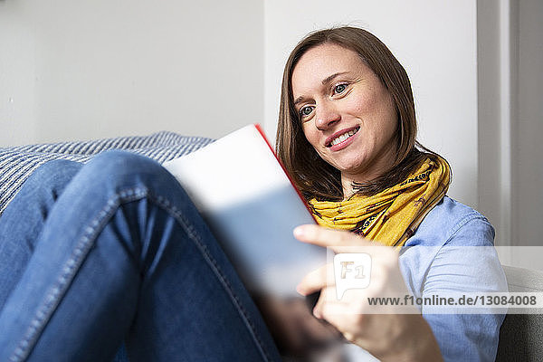 Lächelnde Frau liest Zeitschrift  während sie zu Hause auf dem Sofa sitzt