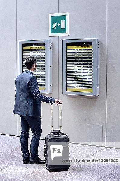 Rückansicht eines Geschäftsmannes  der die Ankunft-Abfahrtstafel liest  während er mit Gepäck in der U-Bahn-Station steht