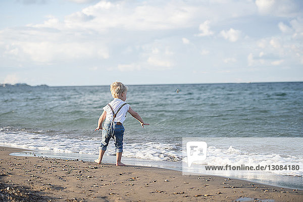 Rear view of baby boy throwing stone in sea while standing at beach against cloudy sky