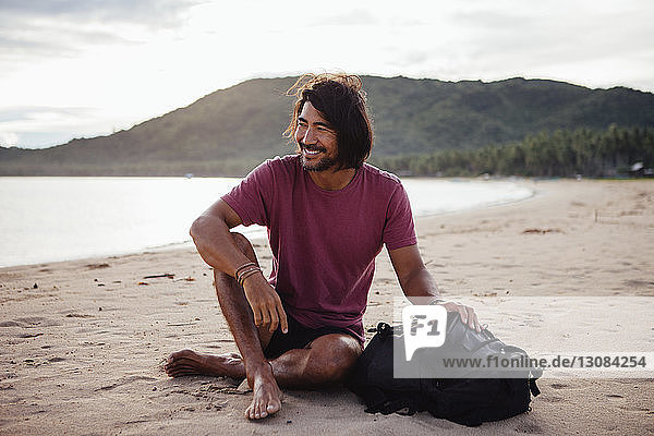 Lächelnder Mann mit Rucksack sitzt am Strand vor bewölktem Himmel
