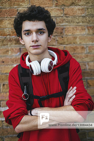 Portrait of confident teenage boy with backpack and arms crossed standing against brick wall