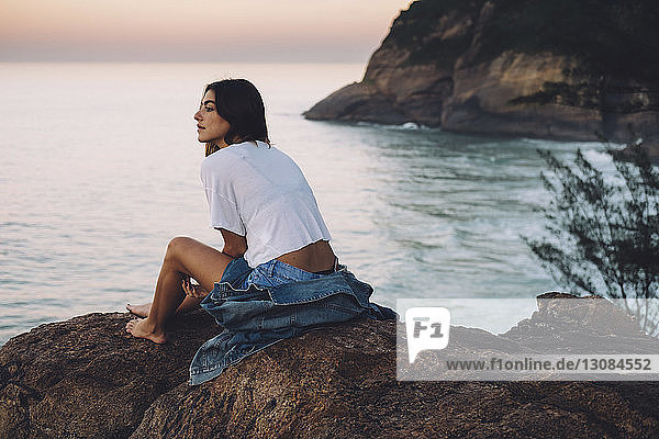 Woman looking at sea while sitting on rock formation against sky during sunset