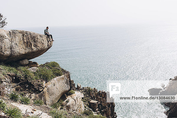 Mid distance view of hiker sitting on cliff while looking at sea
