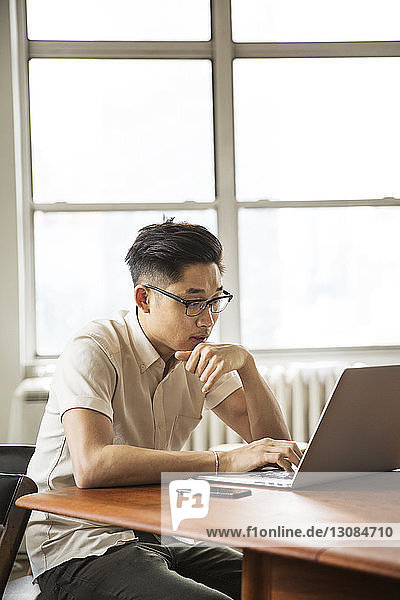 Geschäftsmann mit Laptop  während er in einem hell erleuchteten Kreativbüro am Tisch am Fenster sitzt