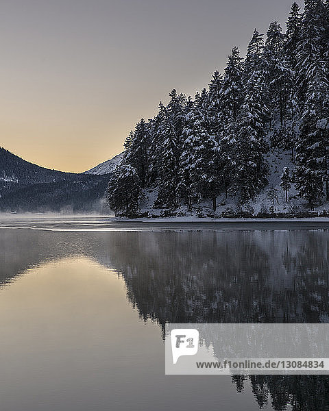 Landschaftliche Ansicht eines ruhigen Sees mit schneebedeckten Bäumen gegen den Himmel bei Sonnenuntergang