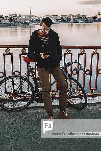 Männlicher Radfahrer benutzt Smartphone  während er bei Sonnenuntergang mit dem Fahrrad auf der Brücke über den Fluss in der Stadt steht