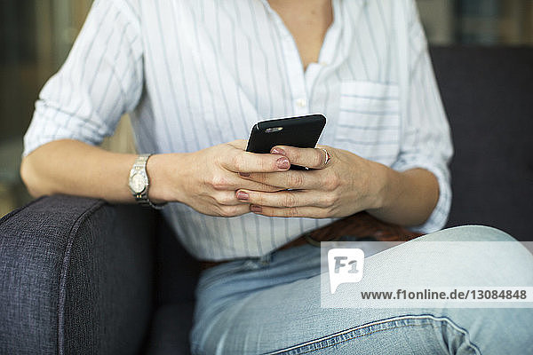 Mittelsektion einer Geschäftsfrau  die ein Mobiltelefon benutzt  während sie im Büro auf dem Sofa sitzt