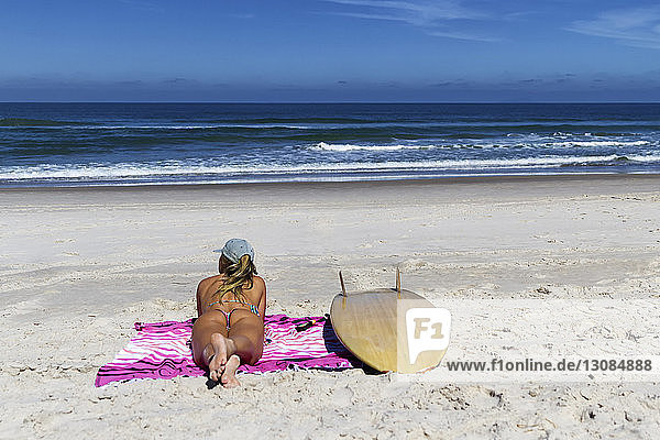 Rückansicht einer Frau im Bikini  die am Strand am Surfbrett auf einer Decke vor blauem Himmel am sonnigen Tag liegt