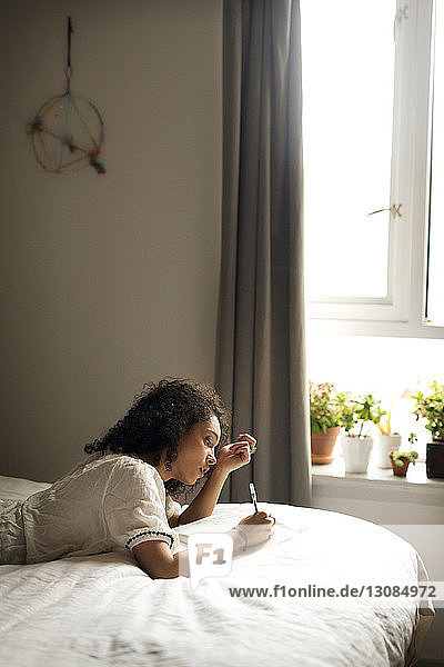 Side view of woman writing on book while relaxing in bedroom