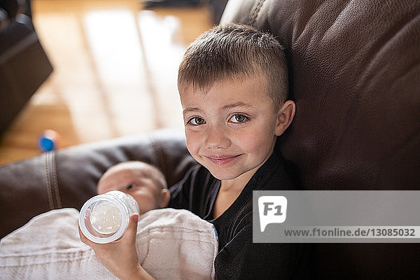 Porträt eines Jungen  der zu Hause auf dem Sofa sitzend seinem Bruder aus der Babyflasche Milch gibt