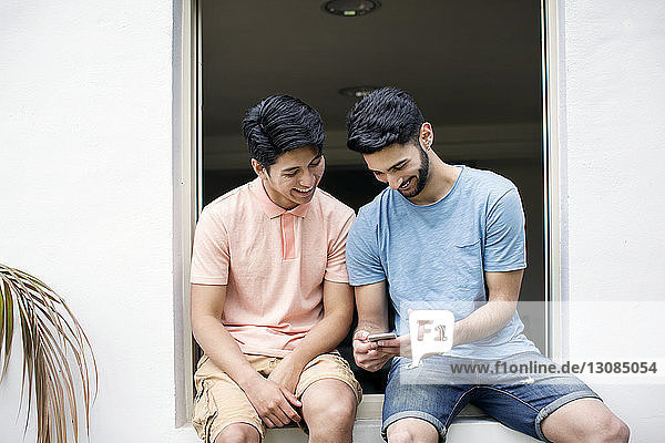 Happy male friends using smart phone while sitting on window