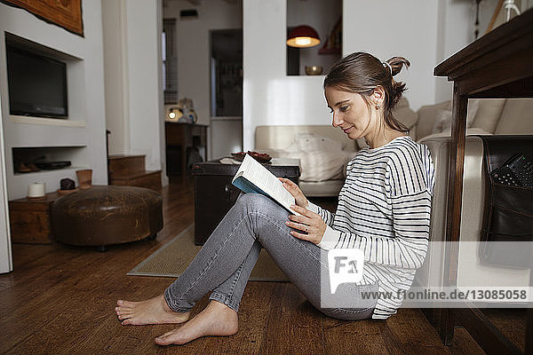Frau liest Buch  während sie zu Hause auf einem Parkettboden sitzt