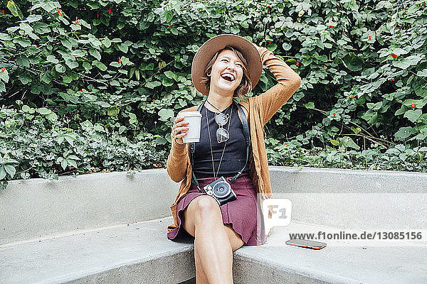 Fröhliche Frau hält Einwegbecher in der Hand  während sie im Park auf einer Bank sitzt