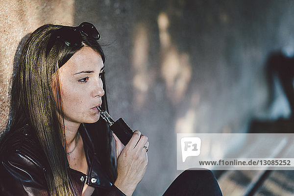 Young woman smoking electronic cigarette while sitting against wall on footpath