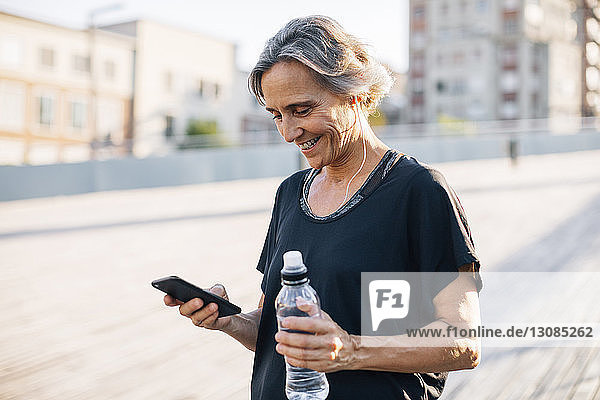 Glückliche Frau schaut auf Smartphone  während sie eine Wasserflasche hält