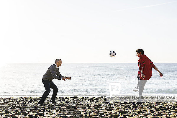 Playful son kicking soccer ball while father defending at beach against clear sky