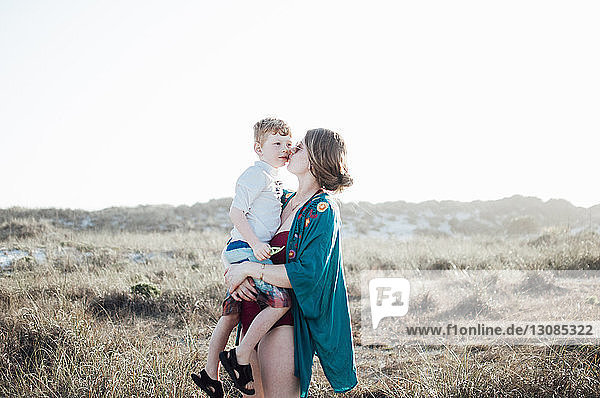 Mutter küsst Sohn  während sie bei strahlendem Sonnenschein auf Grasfeld vor klarem Himmel steht