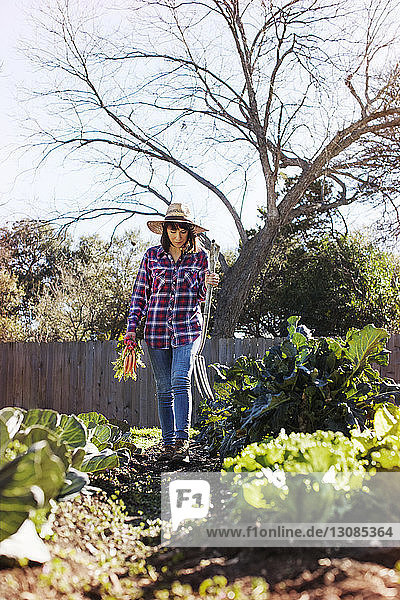 Female farmer holding vegetables and gardening fork while walking at organic farm