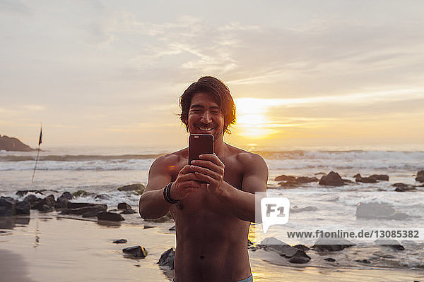 Mann hält Smartphone in der Hand  während er bei Sonnenuntergang am Strand steht