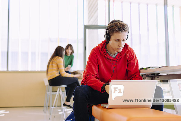Mann benutzt Laptop  während Freundinnen im Hintergrund in der Bibliothek lernen