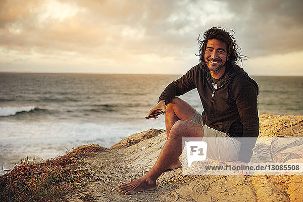 Porträt eines glücklichen Mannes mit Handy in der Hand  der auf einer Klippe am Meer sitzt