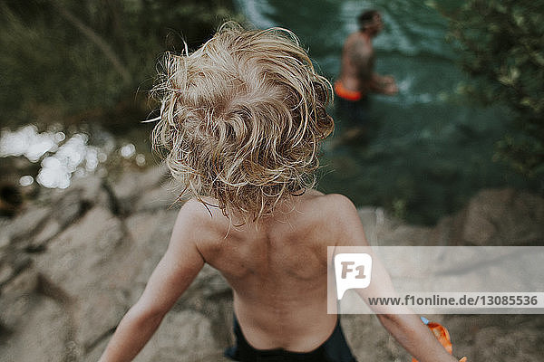 Rückansicht eines Jungen ohne Hemd  der den im Fluss schwimmenden Vater betrachtet