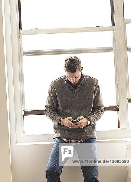 Businessman using mobile phone while leaning on window sill at brightly lit office