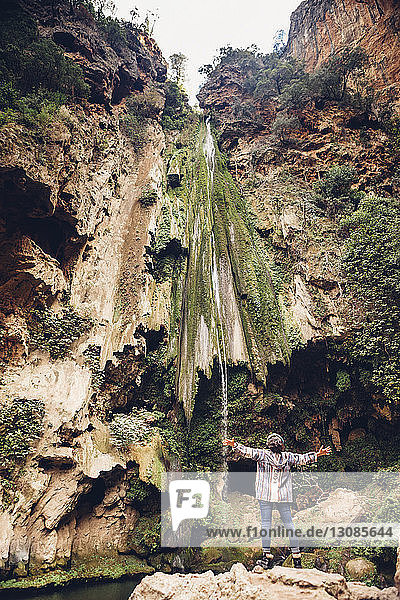 Rückansicht einer Wanderin mit ausgestreckten Armen am Wasserfall über einer Felsformation stehend