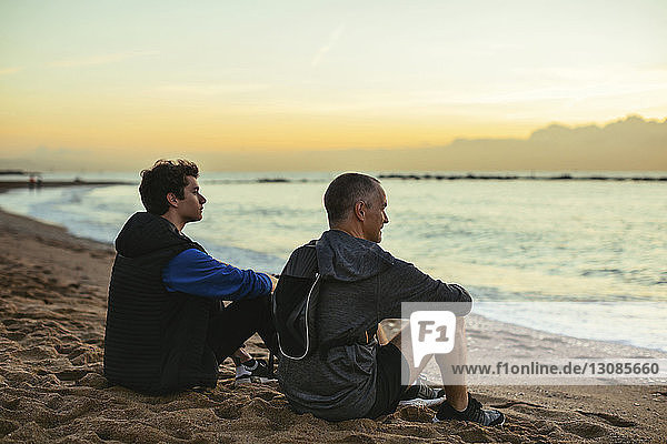 Nachdenklicher Vater und Sohn ruhen sich aus  während sie bei Sonnenuntergang am Strand gegen den Himmel sitzen