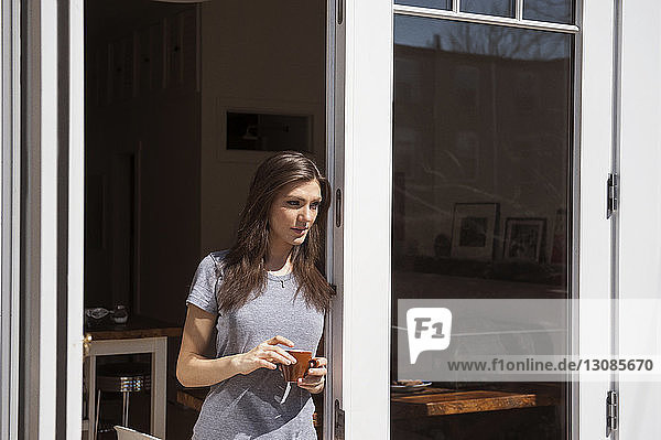 Frontansicht einer Frau  die eine Kaffeetasse hält und am Fenster steht