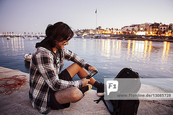 Smiling backpacker looking at phone while sitting by sea during sunset