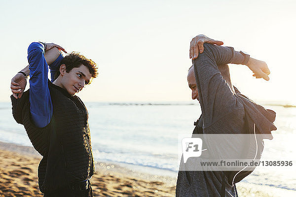 Father and son stretching arms while standing face to face at beach against sky