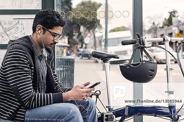 Männlicher Pendler benutzt Smartphone  während er mit dem Fahrrad an der Bushaltestelle sitzt