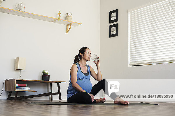 Schwangere Frau trinkt Wasser  während sie zu Hause auf der Gymnastikmatte sitzt