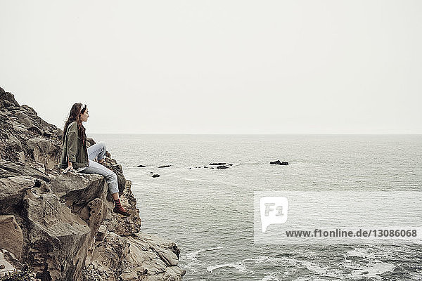 Nachdenkliche Frau sitzt auf einer Klippe am Meer vor klarem Himmel