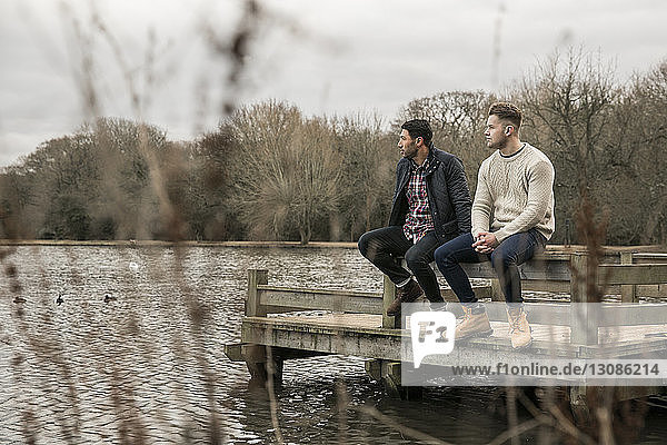 Friends looking away while sitting on pier over lake in Epping Forest
