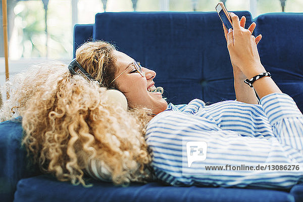 Side view of cheerful woman using smart phone while lying on sofa at home