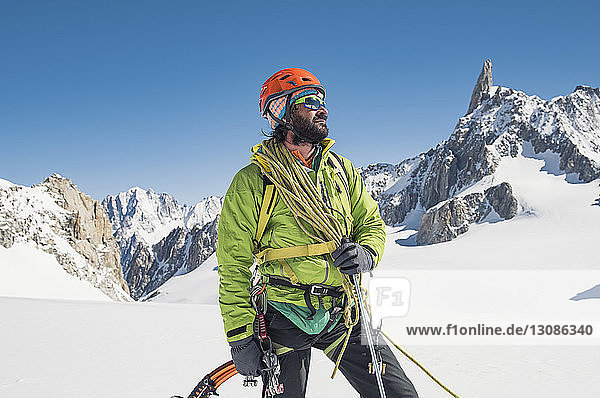 Wanderer mit Kletterausrüstung steht auf schneebedecktem Berg vor strahlend blauem Himmel